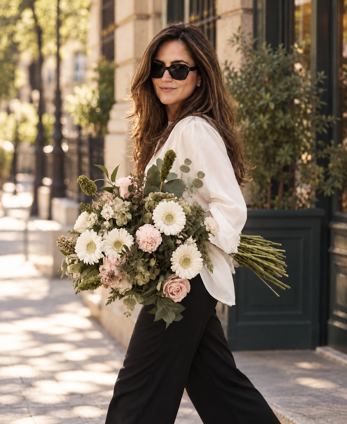 Mujer con camisa blanca y gafas de sol caminando por la calle con un gran ramo de gerberas blancas, rosas y claveles rosa pálido y verdes