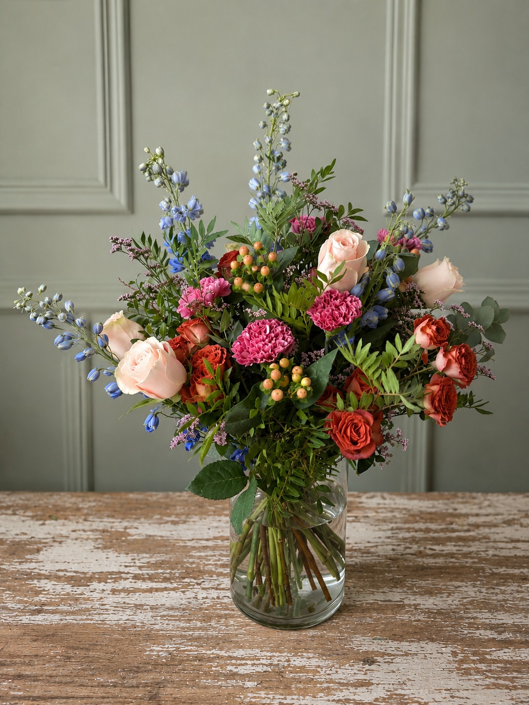 Ramo de flor fresca en jarrón de cristal con rosas rosa pálido y naranjas, claveles fucsia, delphinium azul e hypericum, sobre una mesa de madera desgastada ante una pared verde salvia